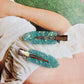 Two green hair clips on a person's arm with a magazine in the background